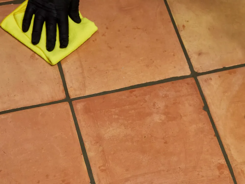 Gloved hand wiping clean terracotta floor tile with a yellow microfiber cloth